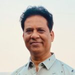 A middle-aged man with short, curly black hair smiles slightly while wearing a light gray collared shirt, standing against a pale, blurred background—suggesting a professional in the field of radiology.