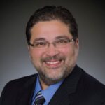 A man with short dark hair, glasses, and a trimmed beard is smiling. He is dressed in a dark suit, blue shirt, and striped tie, posing confidently against a dark gradient background—reflecting professionalism in radiology.