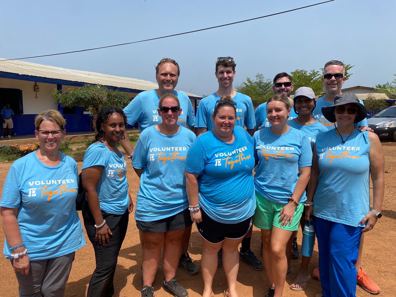 A group of twelve people in blue VOLUNTEER Together T-shirts pose and smile outdoors on a sunny day, proudly representing their RP team as they stand on reddish dirt with trees and buildings in the background.
