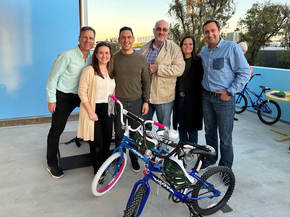 Six adults stand together, smiling on a sunny patio with trees in the background. Two children's bicycles, one pink and one blue, sit in front of them—perfect for an afternoon RP adventure under pleasant skies.