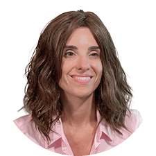 Woman with wavy brown hair wearing a light pink collared shirt, smiling against a plain white background.
