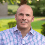 A man with a shaved head, wearing a pastel plaid button-up shirt, smiles outdoors with green grass and trees in the blurred background—one of our celebrated value award winners.