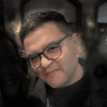 A man with short dark hair and glasses, wearing a black shirt and jacket, smiles slightly while looking at the camera in a dimly lit indoor setting, resembling one of the value award winners at an exclusive event.