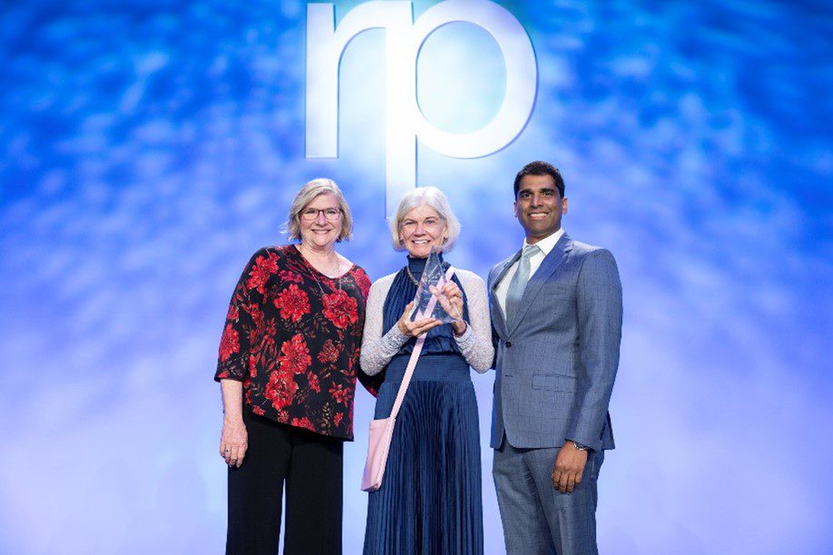Three people stand smiling on stage in front of a blue backdrop with the letters “rp.” The person in the center holds a clear award for best practice recommendation. The person on the right wears a suit; the others wear patterned and blue outfits.